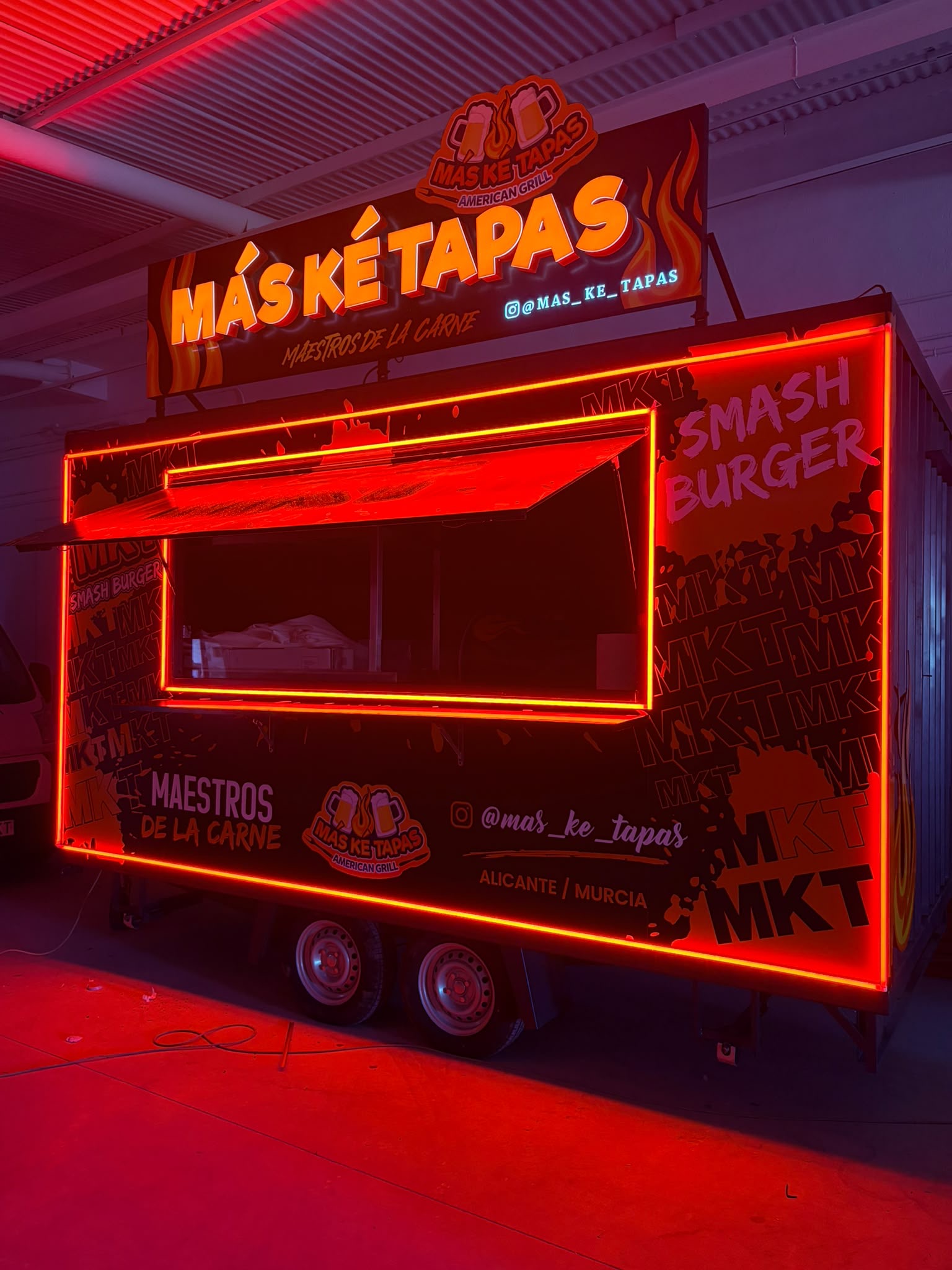 Foodtruck de MÁS KE TAPAS American Grill iluminada con neón rojo, con el rótulo Maestros de la carne y el logo de la marca