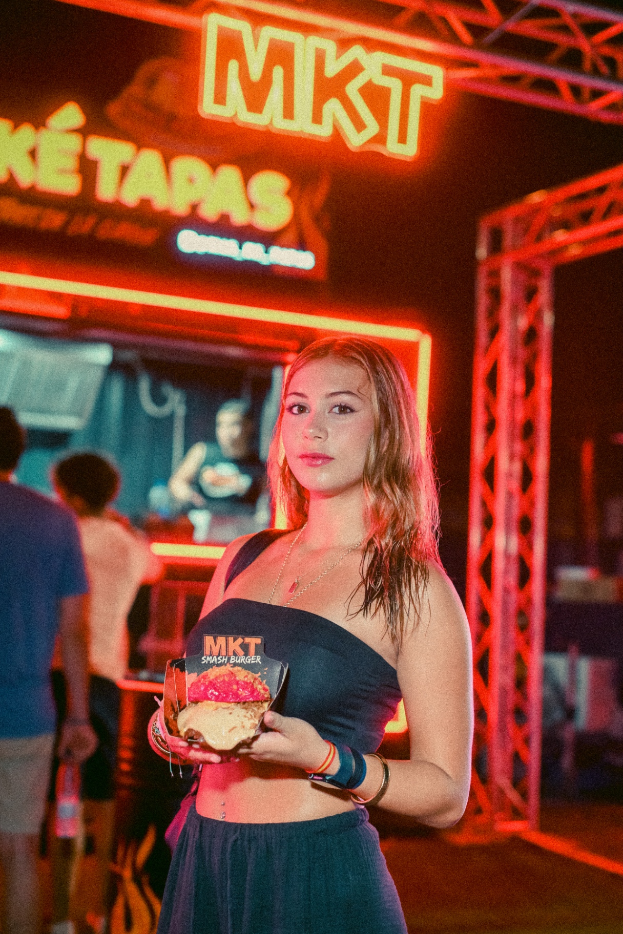 Clienta sosteniendo una smash burger MKT frente al rótulo luminoso MÁS KÉ TAPAS