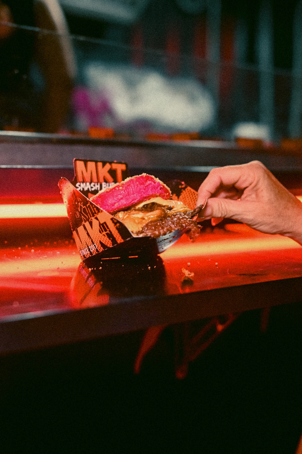 Detalle de una smash burger MKT con pan rosa y queso fundido sobre la barra iluminada